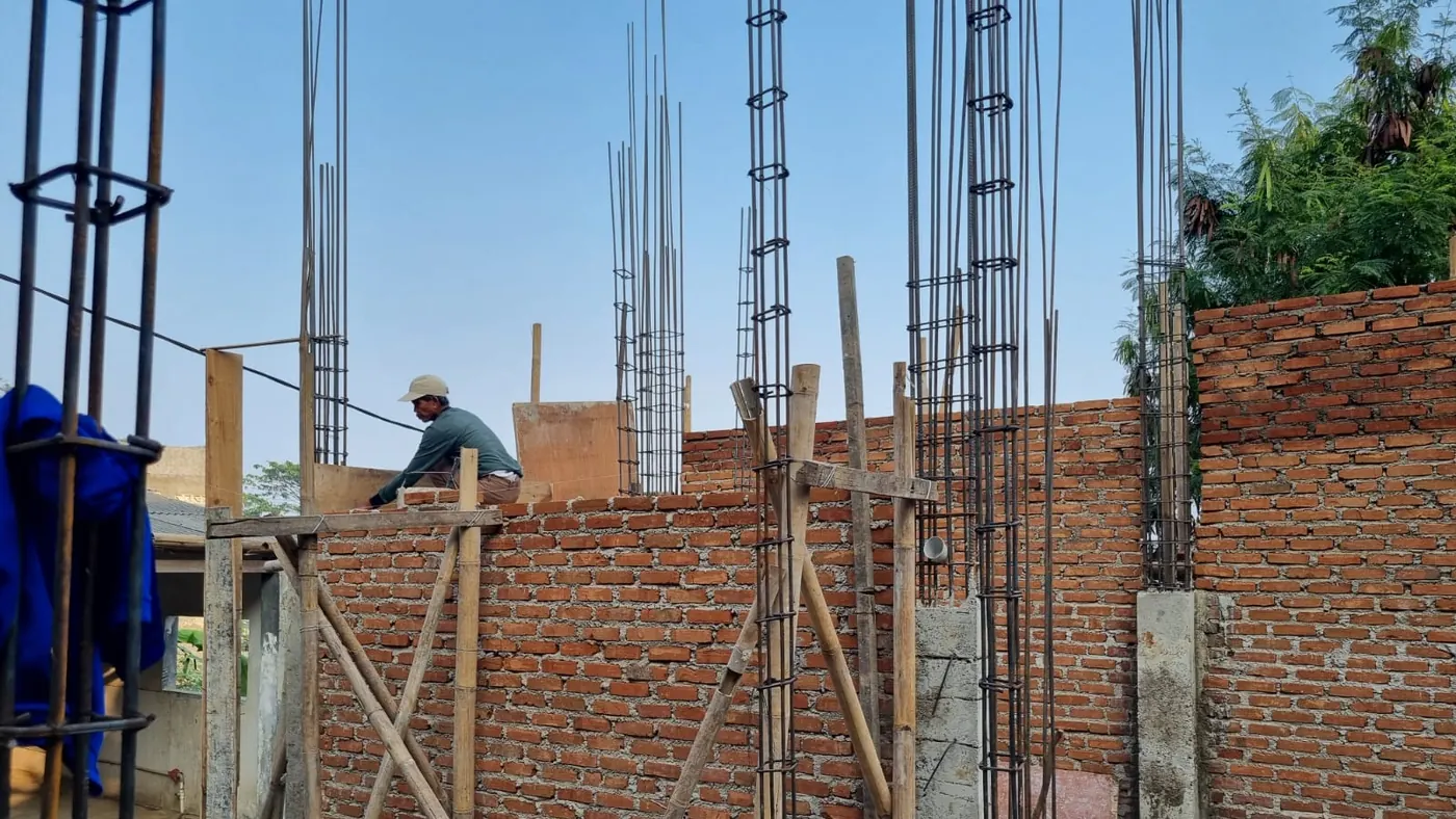 Seorang pekerja memasang bata merah pada dinding Rumah Tahfidz yang setengah jadi, dengan kolom beton dan perancah bambu