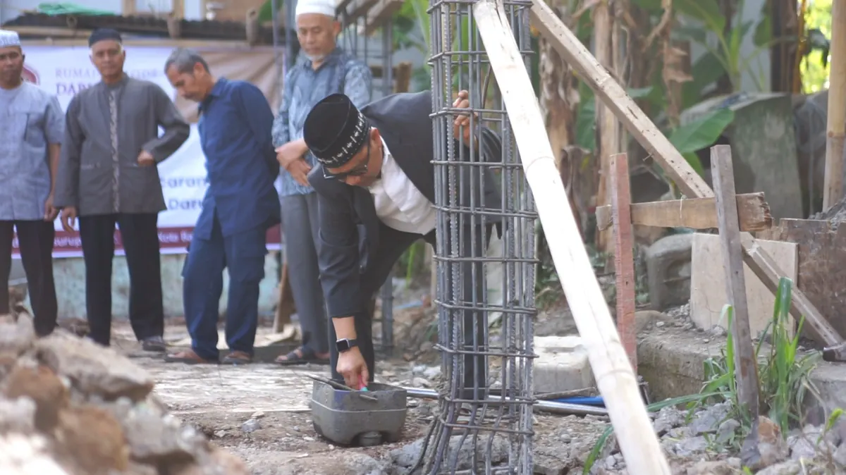 Pembina Yayasan berpeci dan berjas membungkuk memakai cetok dalam acara peletakan batu pertama, disaksikan para tokoh setempat di lokasi pembangunan