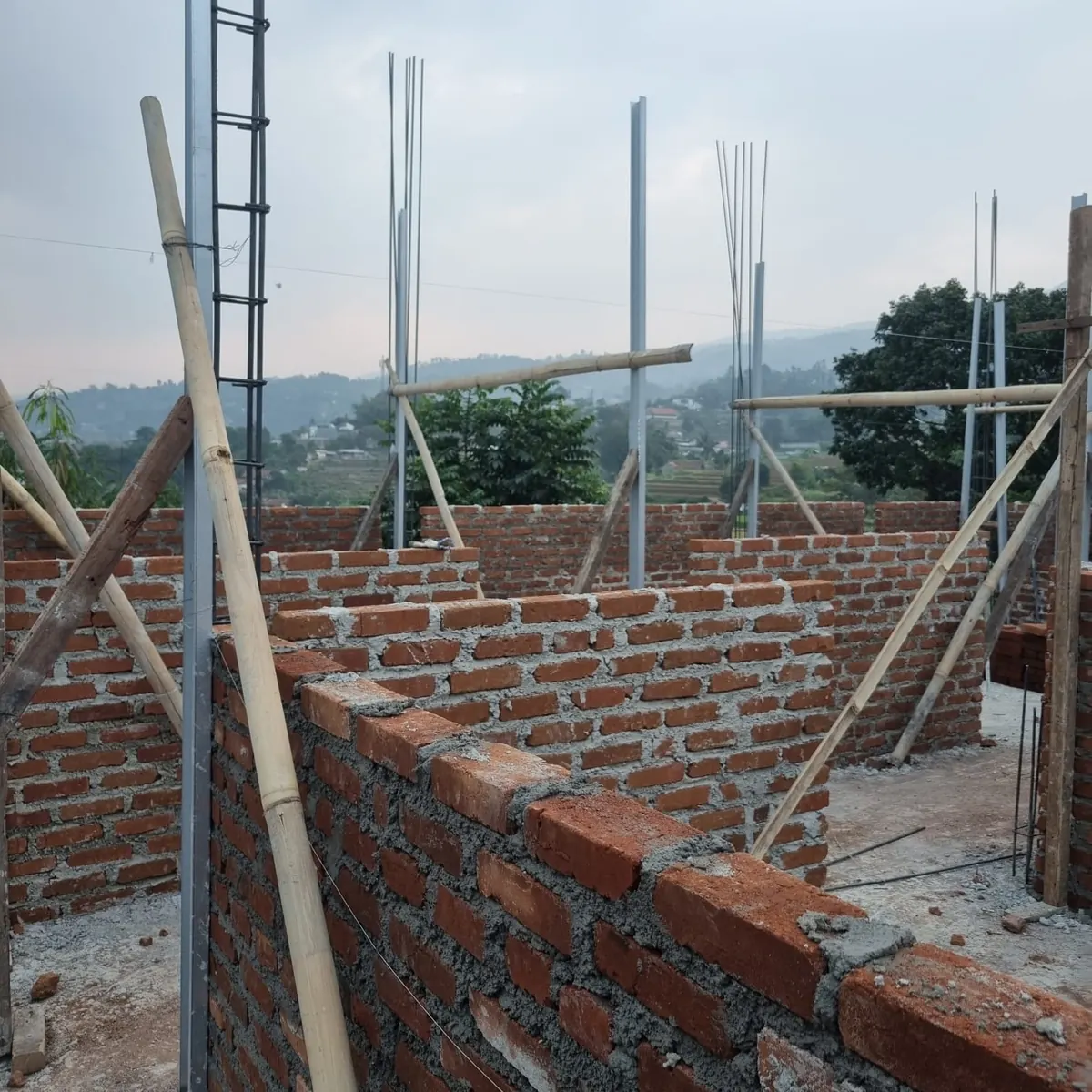 Dinding bata merah sudah tersusun tinggi membentuk sekat ruangan, dengan besi tulangan menjulang dan bambu penopang